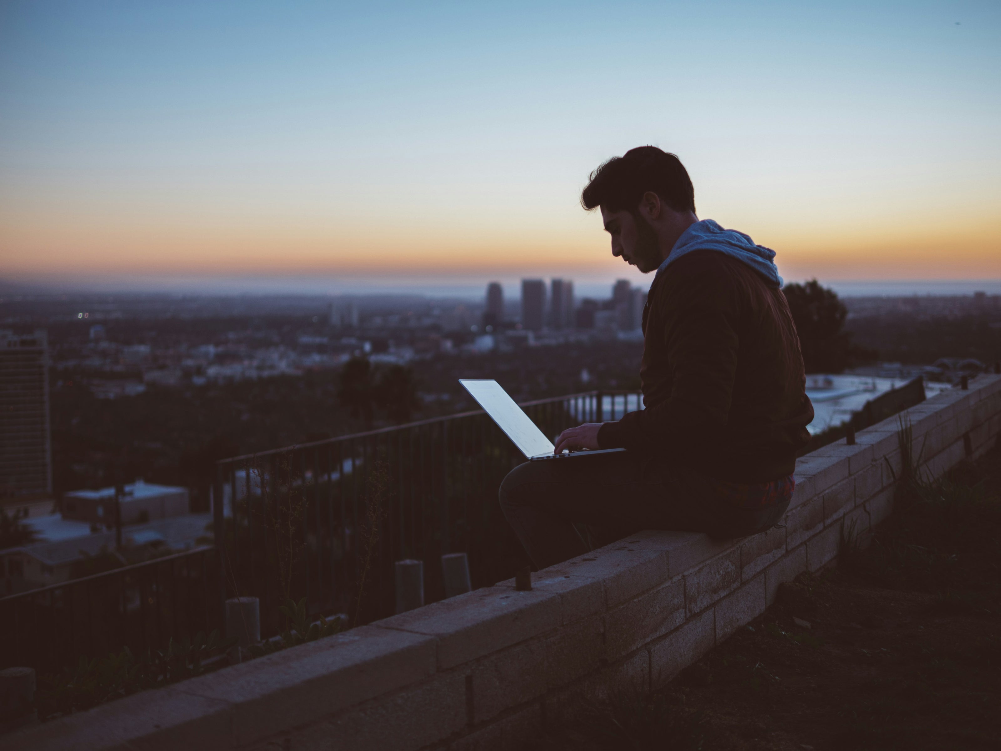 Person sitting on a ledge with a laptop at sunset, overlooking a cityscape.